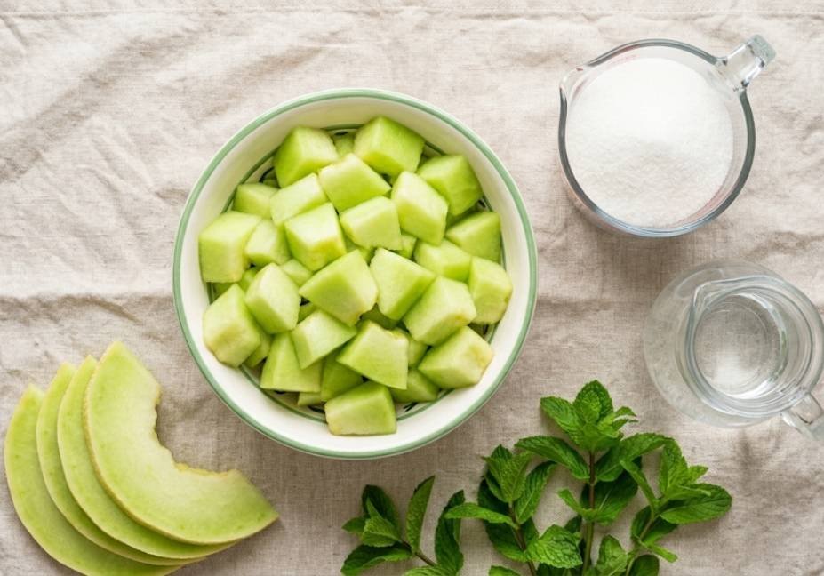 Flat lay view of honeydew syrup ingredients including chopped melon chunks in a bowl, white sugar, water, melon slices, and fresh mint sprigs."