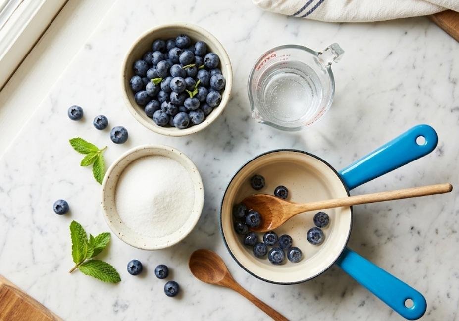 Overhead view of ingredients for blueberry simple syrup, featuring fresh blueberries, white sugar, water in a measuring cup, and a blue enamel saucepan on a marble countertop.